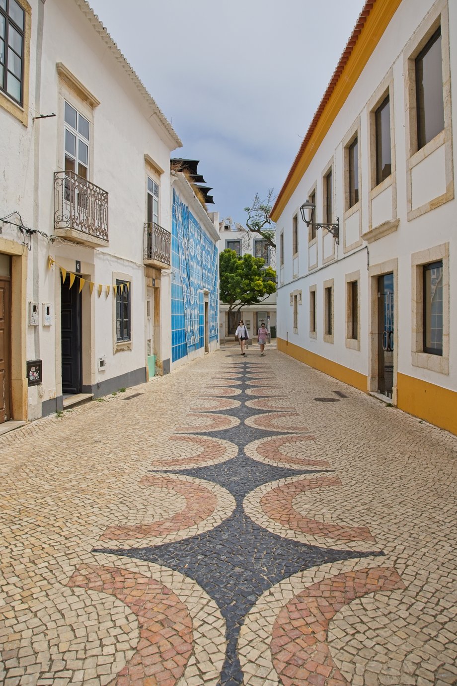 A narrow cobblestone street in a traditional Portuguese town, featuring white buildings with stone window frames and a yellow-painted roofline on the right side. The street is paved with an intricate mosaic cobblestone pattern in blue, white, and terracotta colors forming wavy geometric designs. On the left, a building displays an ornate blue-and-white tiled mural wall. Ornate iron balconies and a dark doorway with yellow bunting flags are visible. Two people are walking down the street toward the camera, and a green tree stands at the far end where the alley opens up under an overcast sky.